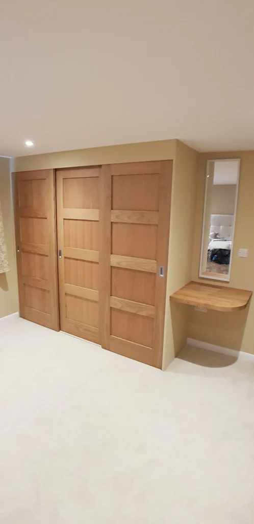Bespoke floor-to-ceiling fitted oak wardrobe in a modern bedroom, featuring three full-height doors with natural wood grain and precision-fit cabinetry in Ninfield, East Sussex.