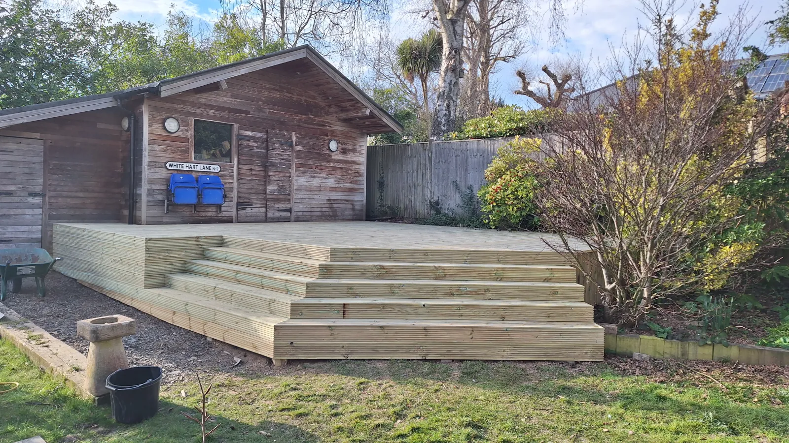 Raised timber decking installation on a sloping garden, featuring structural pressure-treated framing, vertical support posts, and non-slip wooden deck boards.