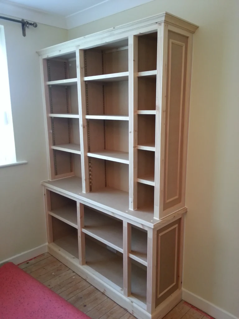 The finished, unpainted library cabinet unit featuring fluted pilasters, adjustable shelving, and a decorative crown cornice.
