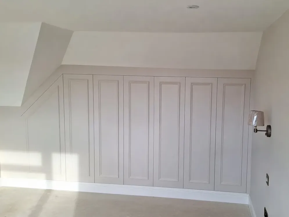 Bespoke fitted wardrobes with traditional panelled doors, custom-built to fit a sloped attic ceiling and finished in a soft grey.