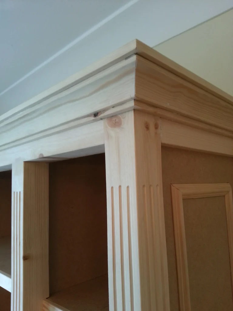 Close-up of the top corner of the cabinet showing the intricate mitred cornice and the vertical fluting on the timber frame.