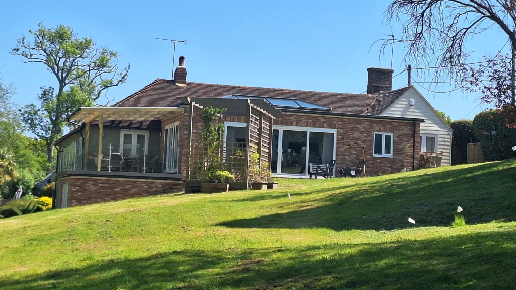 A wide-angle garden view of a brick house featuring a newly constructed timber pergola on a raised terrace overlooking a sloped lawn.
