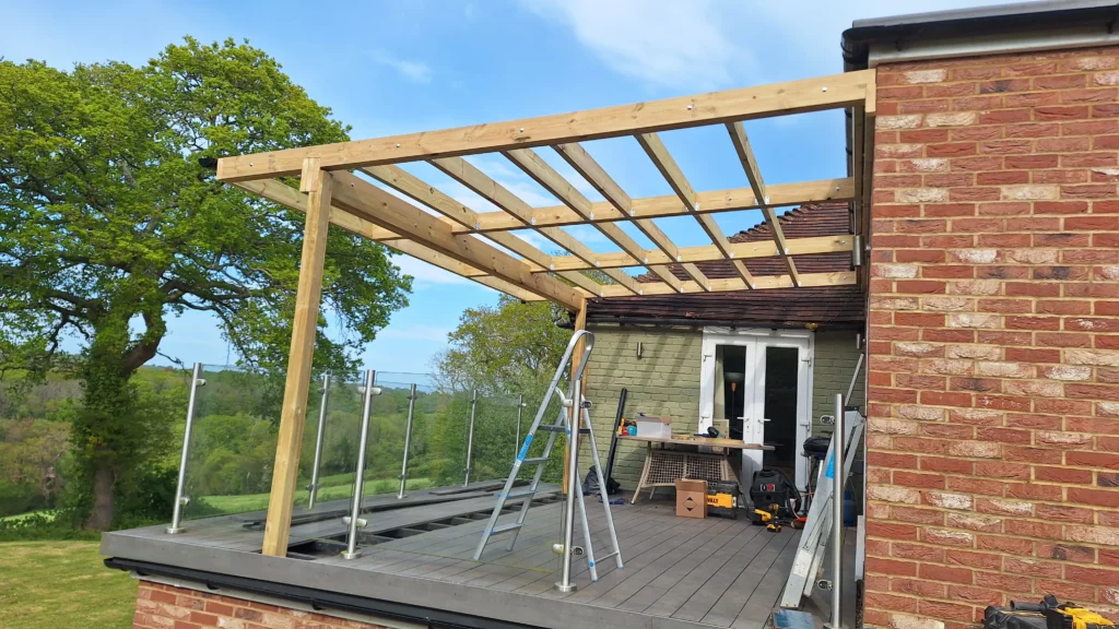 A side view of a bespoke timber pergola built over an elevated balcony with a glass balustrade, showing the structural posts and roof pitch.