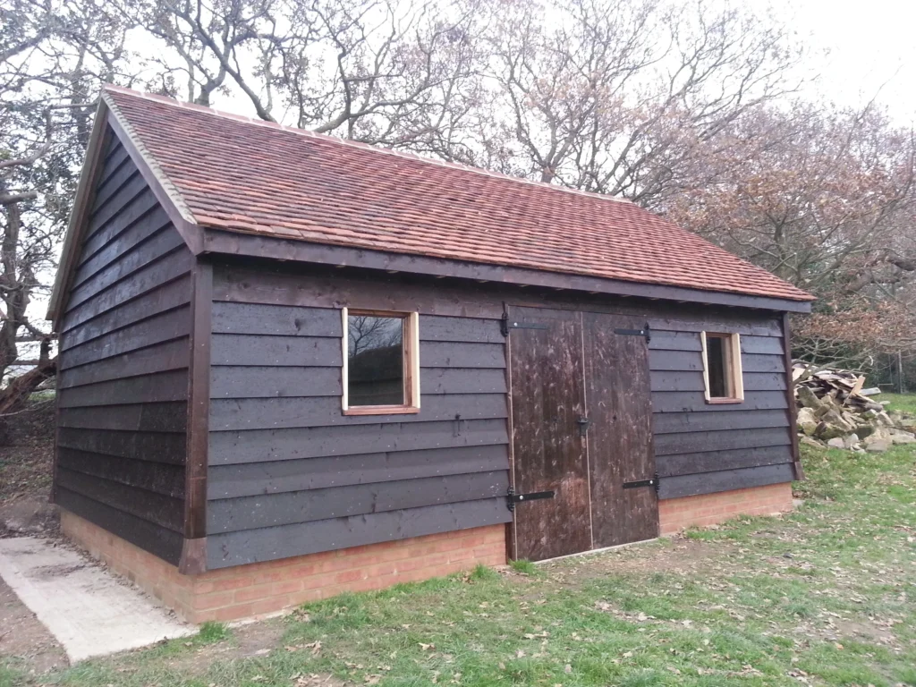 Completed traditional timber-framed garden outbuilding featuring dark weatherboard cladding, a clay tile roof, and a red brick foundation plinth.
